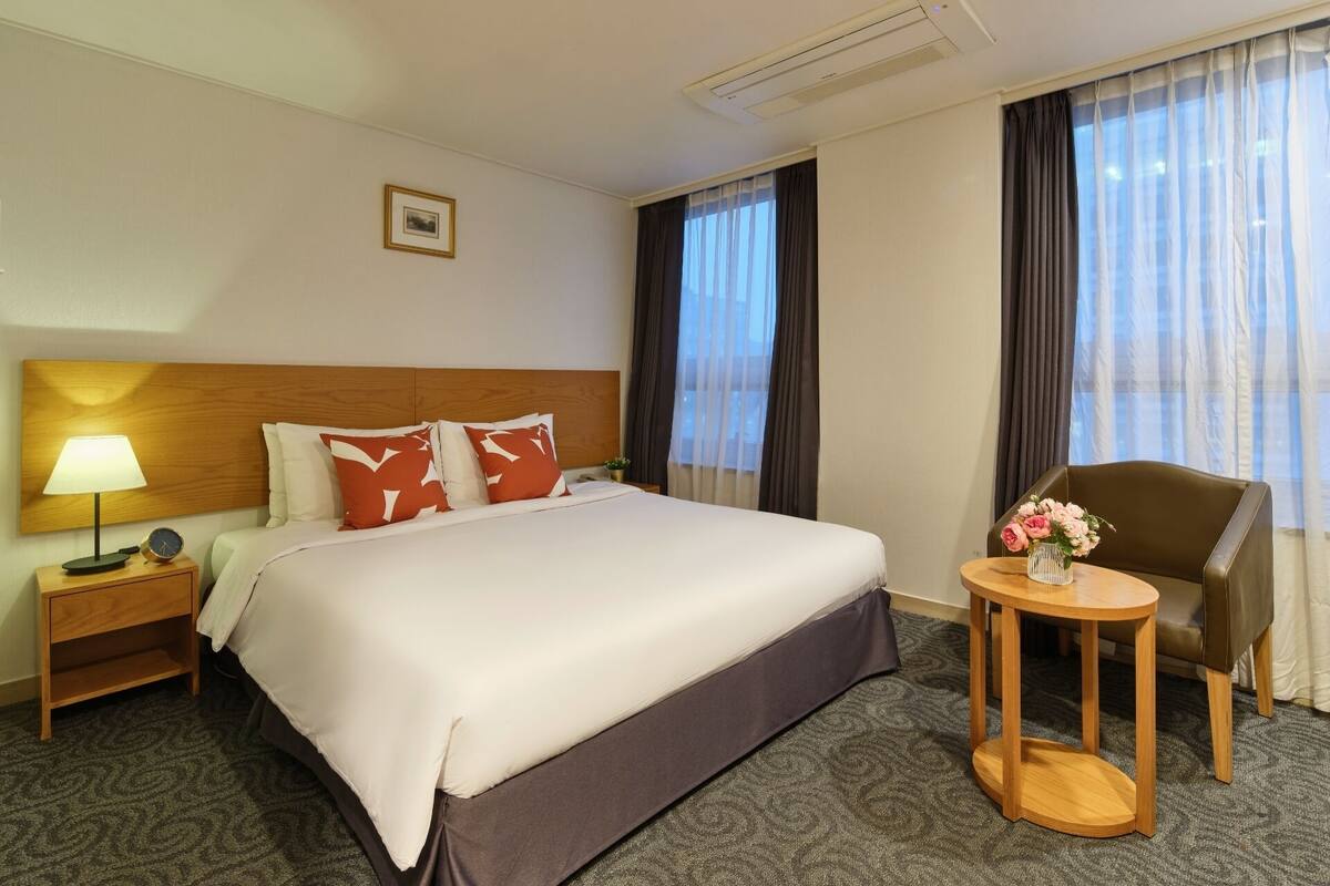 A hotel room with a large bed, a bedside table with a lamp, a chair, and a small table with flowers.