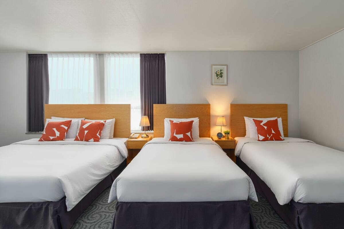 A hotel room with two beds, each with white bedding and red pillows, a wooden headboard, a bedside table with a lamp, and a window with curtains.