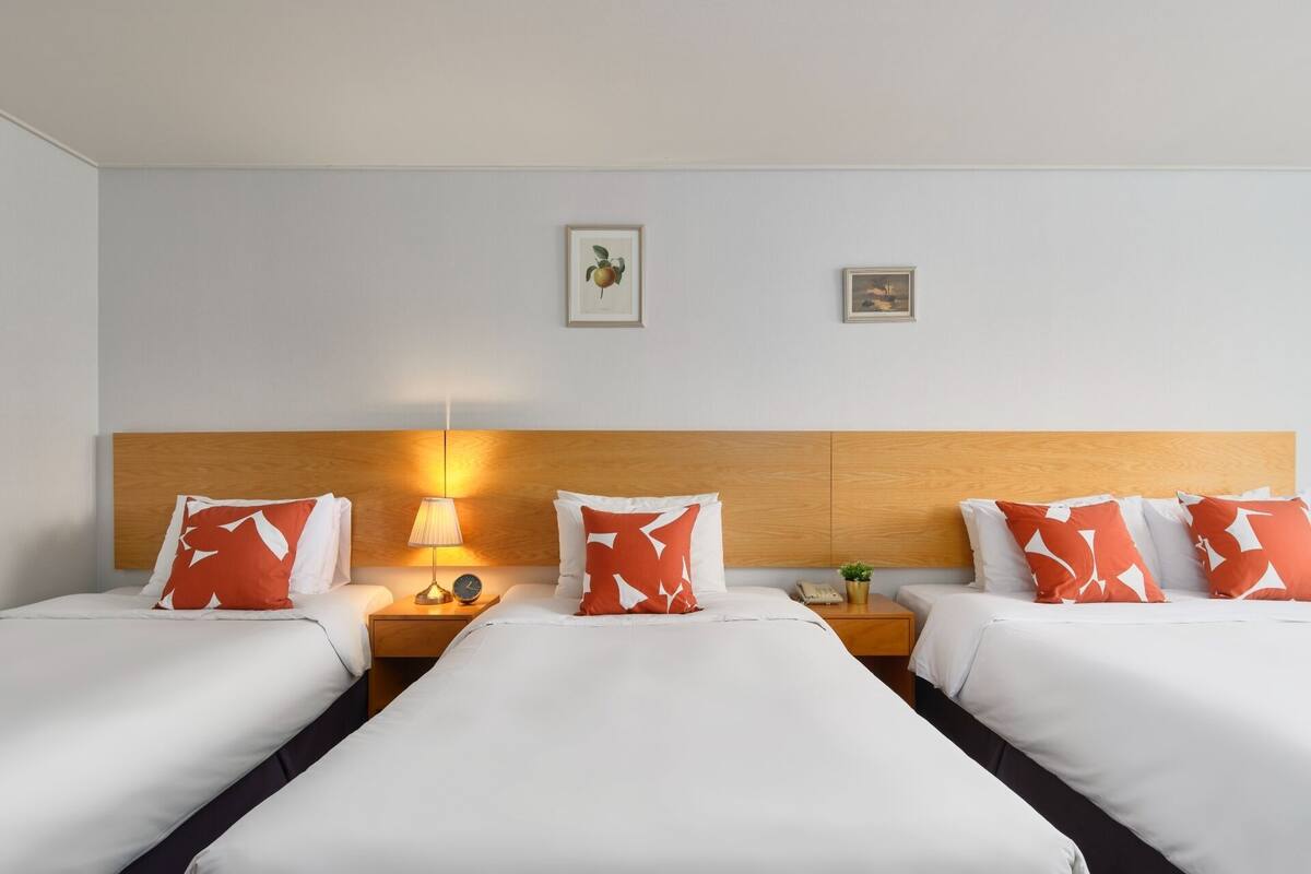 A hotel room with two beds, a wooden headboard, a bedside table with a lamp, and a small clock.