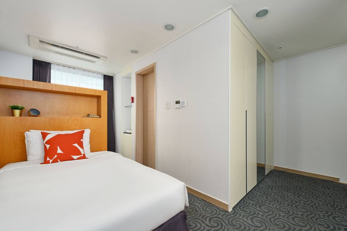 A hotel room with a bed, a wooden headboard, a wall-mounted air conditioner, a thermostat, and a patterned carpet.