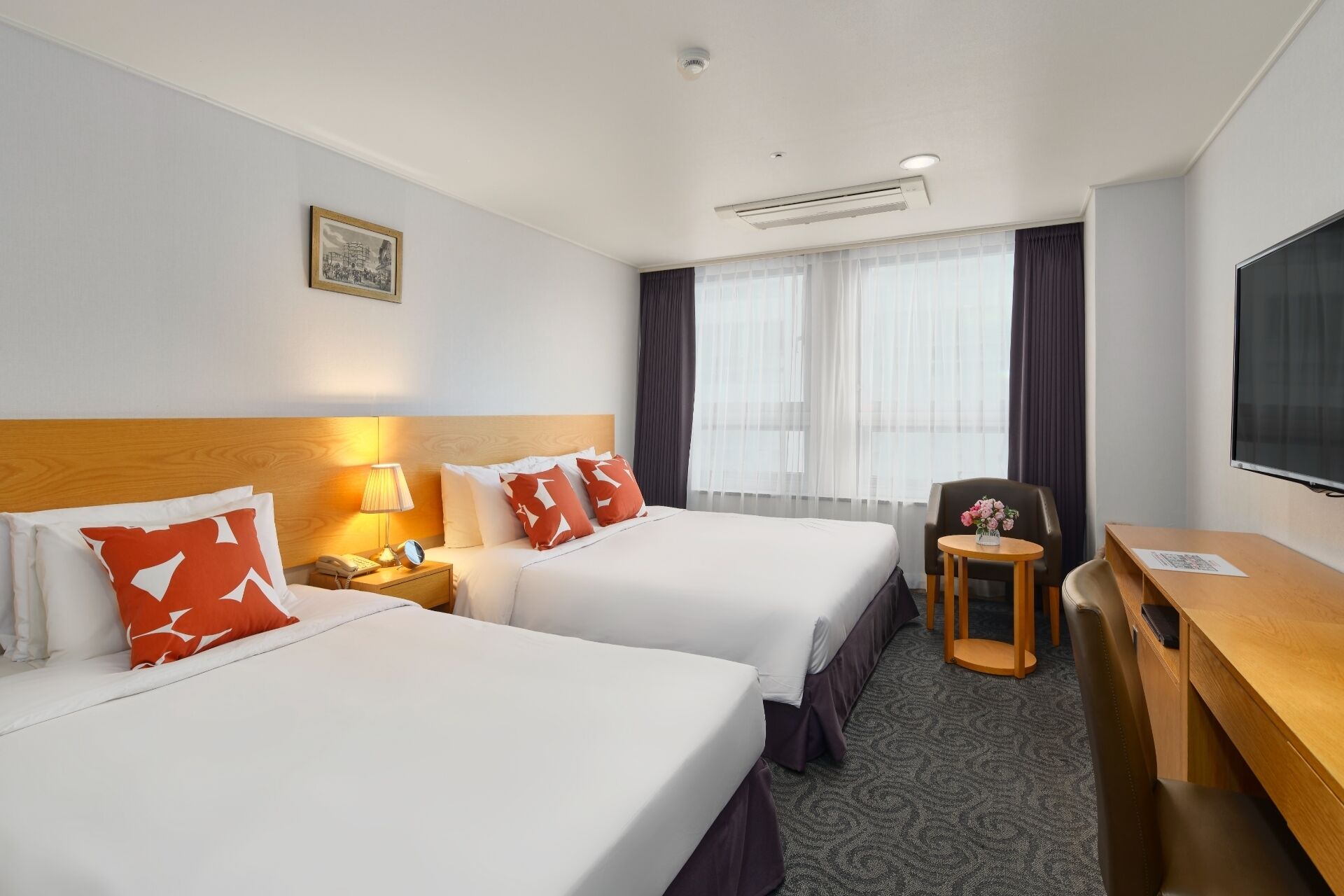 Quarto de hotel com duas camas, uma mesa, uma cadeira, uma televisão e uma janela com cortinas.