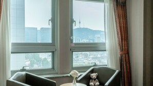 A room with two chairs, a small table with a vase of flowers, and a teddy bear. Large windows offer a view of a cityscape with buildings and a tower. - Hotel Atrium Seoul Jongno (Seoul)