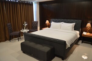 A bedroom with a large bed, two bedside tables with lamps, a round side table with a vase, and a dark upholstered bench.