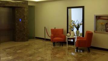 Um hall com piso de mármore, duas cadeiras laranja, uma pequena mesa com um vaso e um espelho grande na parede.