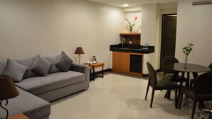 Een moderne woonkamer met een grijze bank, een klein houten bijzettafeltje, een lamp, een eethoek met een ronde tafel en stoelen, een keuken met een zwart aanrecht en houten kastjes, en een deur die naar een andere kamer leidt.