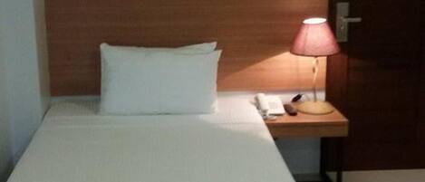 Un lit simple avec un oreiller blanc et une tête de lit en bois. On aperçoit une table de chevet avec une lampe et un téléphone.