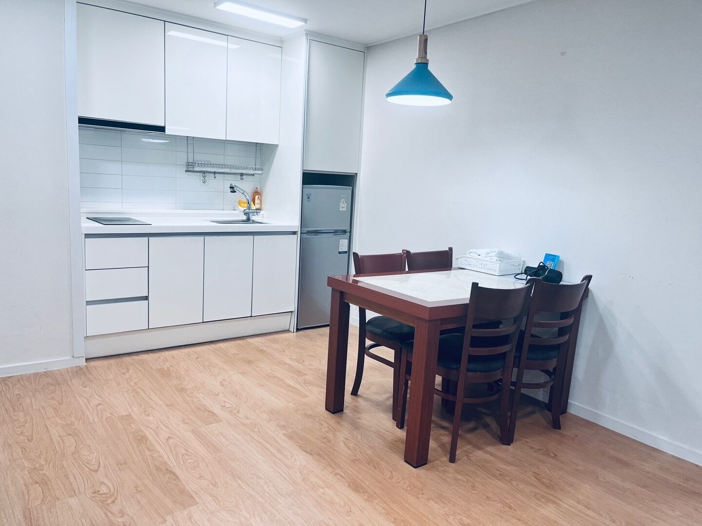 Una cocina moderna con armarios blancos, un lavamanos de acero inoxidable y un horno empotrado. Hay una mesa de comedor con sillas y una lámpara colgante azul por encima.