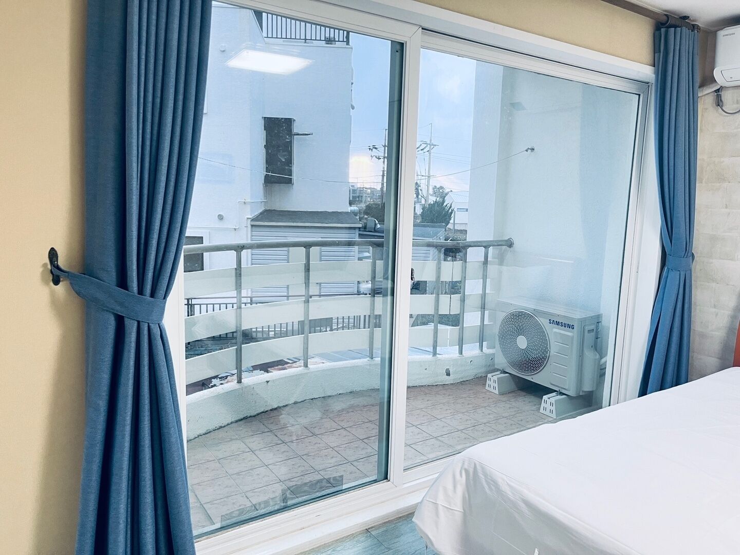 Ein Zimmer mit einer Glasschiebetür, die auf einen Balkon führt, einem Bett mit weißer Bettdecke und einer Samsung-Klimaanlage auf dem Balkon.
