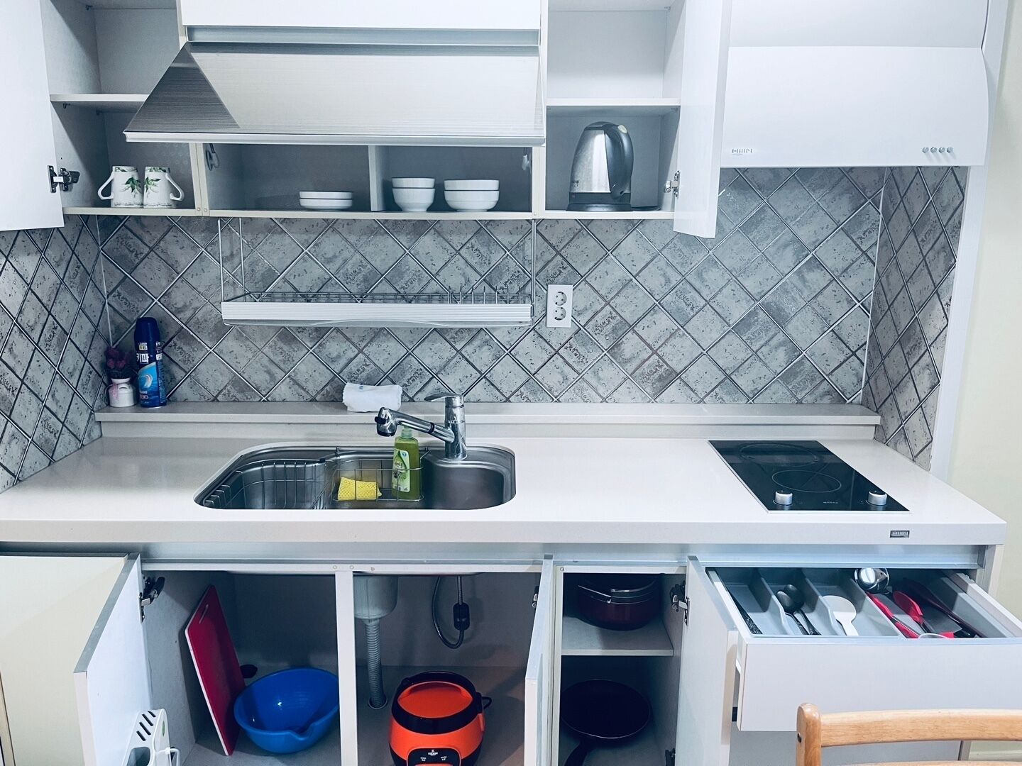 A kitchen with a stainless steel sink, a kettle, and a built-in stove. There are cabinets above the sink and a drawer below.