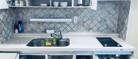 A kitchen with a stainless steel sink, a kettle, and a built-in stove. There are cabinets above the sink and a drawer below.
