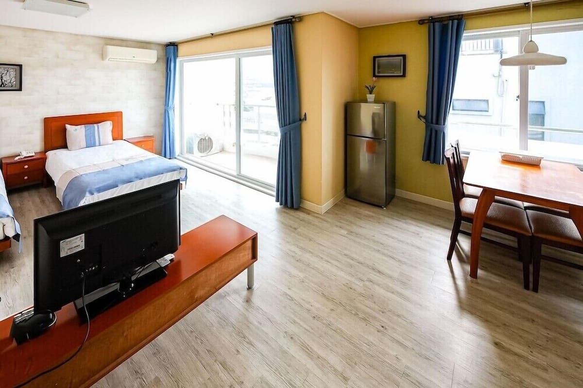 A hotel room with a bed, a TV on a stand, a dining table with chairs, a refrigerator, and a window with curtains.