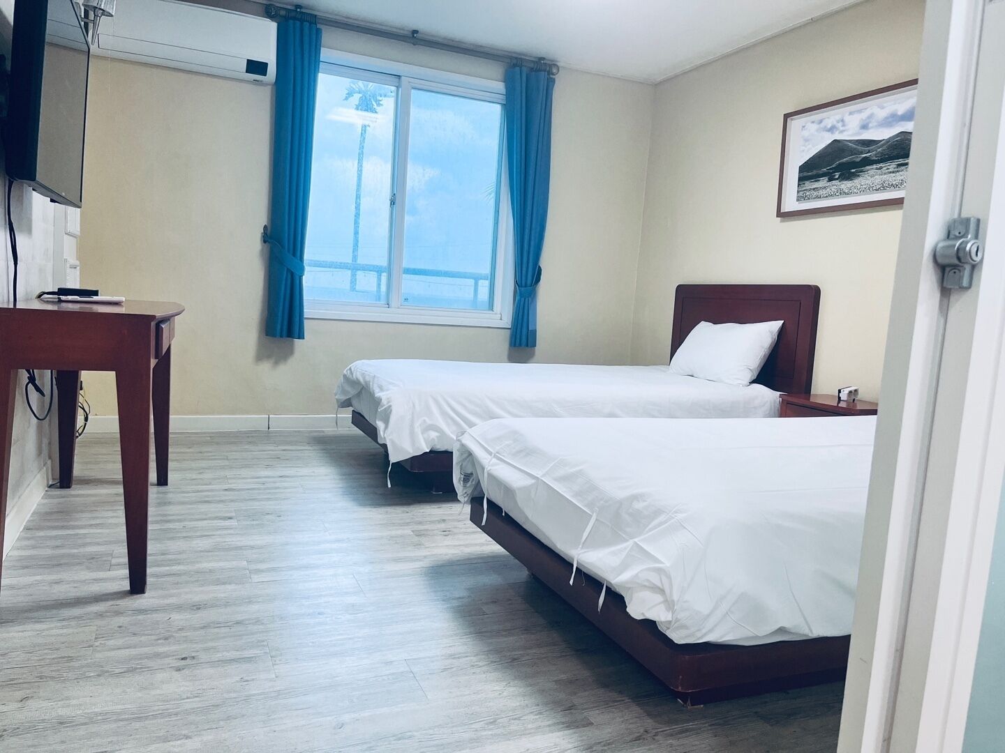 Habitación de hotel con dos camas, un escritorio, un ventanal con cortinas y un cuadro en la pared.