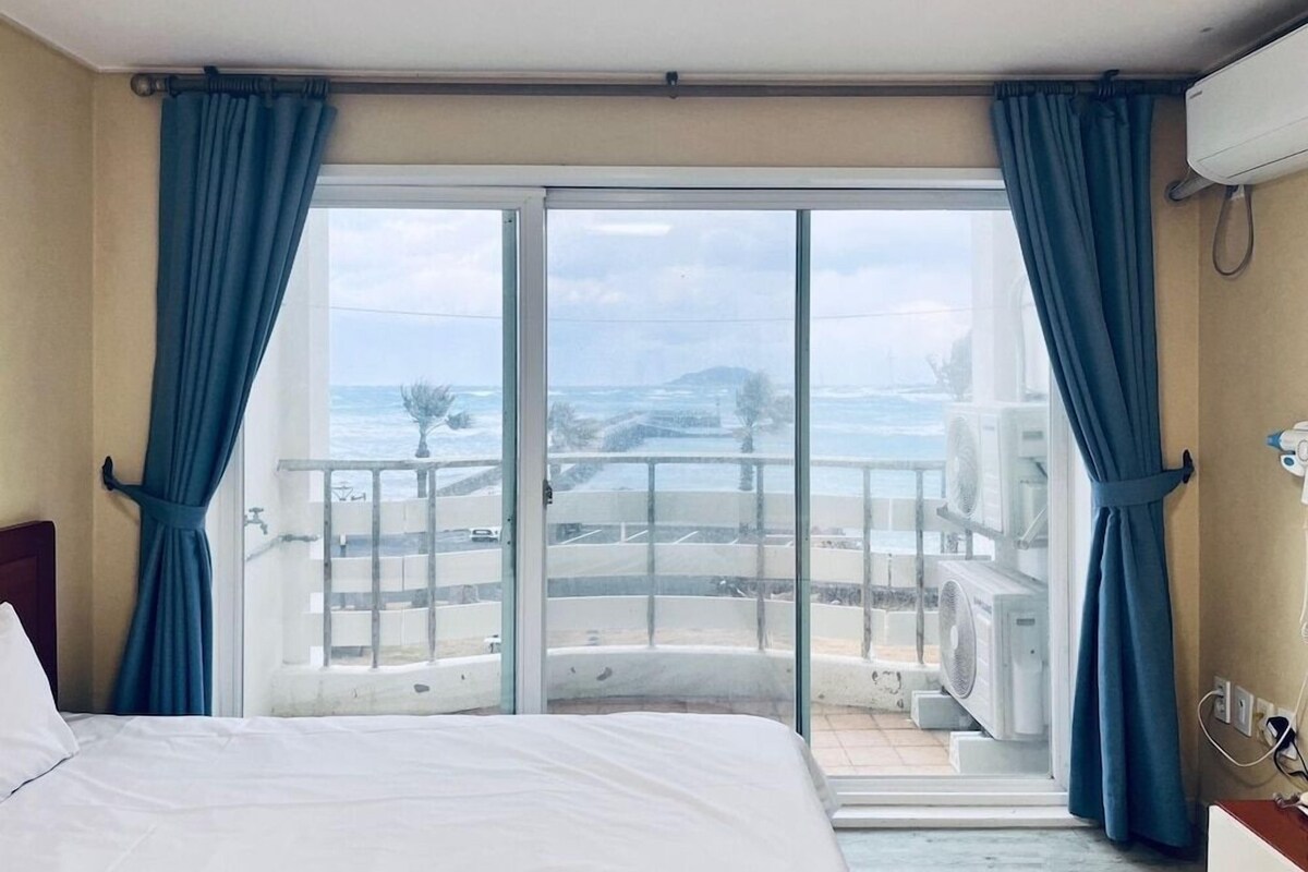 A hotel room with a bed, a view of the sea, and a balcony with a railing.