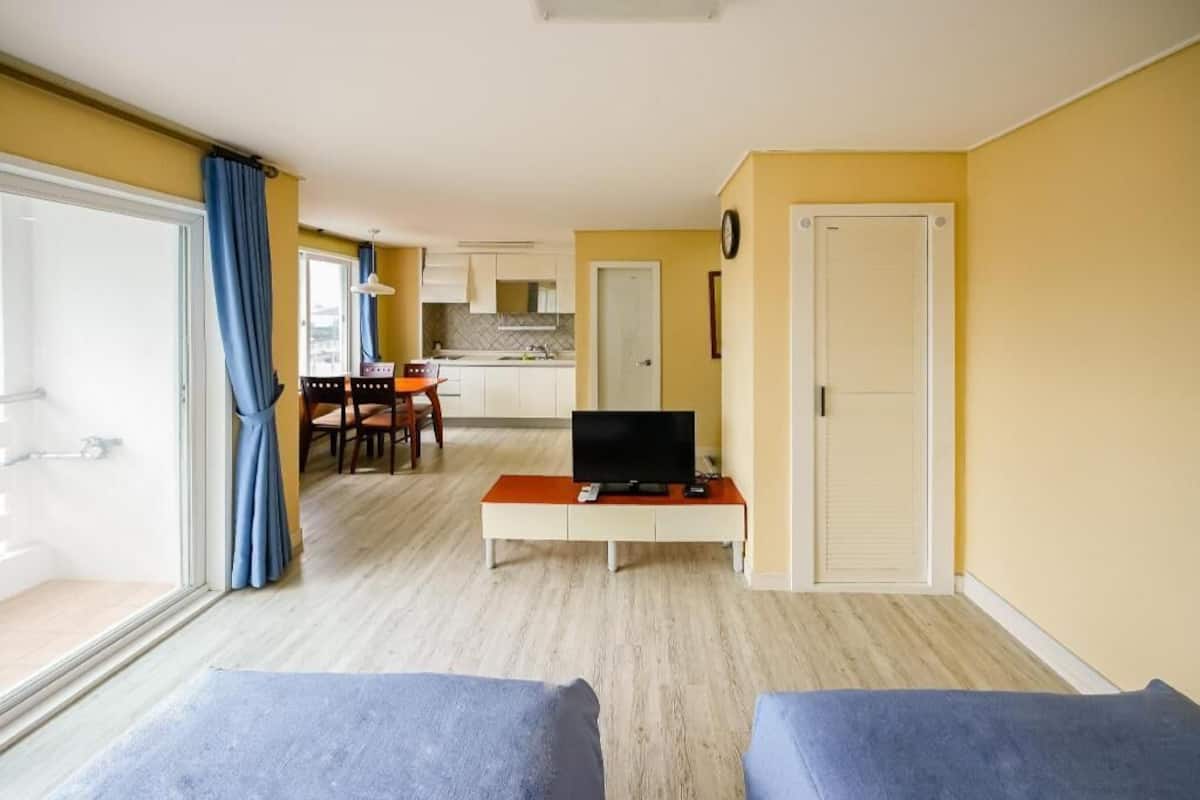 A room with a TV on a stand, a dining table with chairs, a kitchen in the background, and a door.