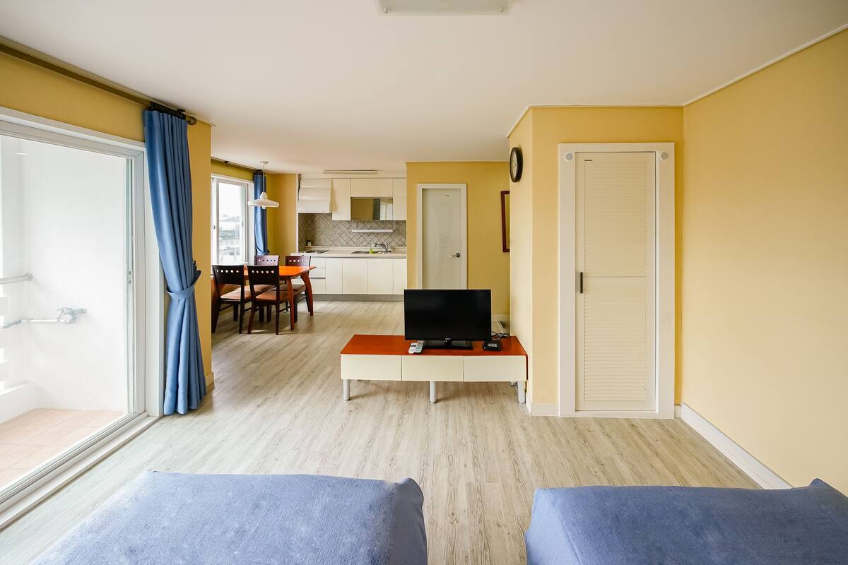 A room with a TV on a small table, a dining area with chairs, and a kitchen in the background.
