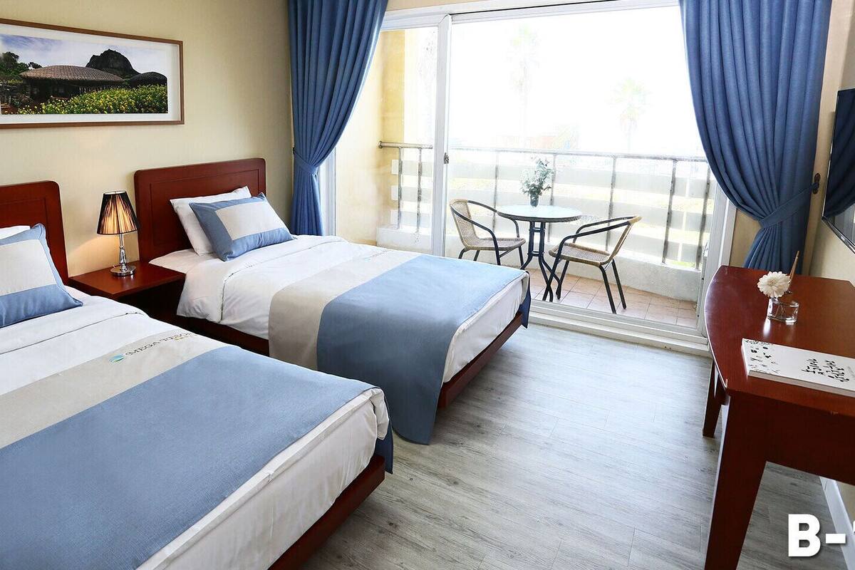 A hotel room with two beds, a small table with chairs, a balcony with a table and chairs, and a framed picture on the wall.