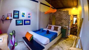 A bedroom with a bed, pillows, a shelf with pictures, a hanging orange object, and a brick wall. - Artist Place Trat (Trat)