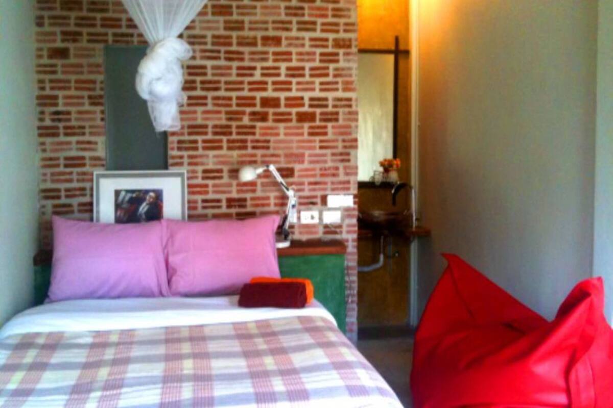 A room with a brick wall, a bed with a plaid bedspread, a nightlight, and a red chair.