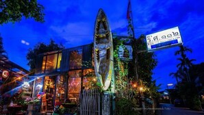 A building with a large, illuminated sculpture in front, a signboard, and a storefront with lights. - Artist Place Trat (Trat)