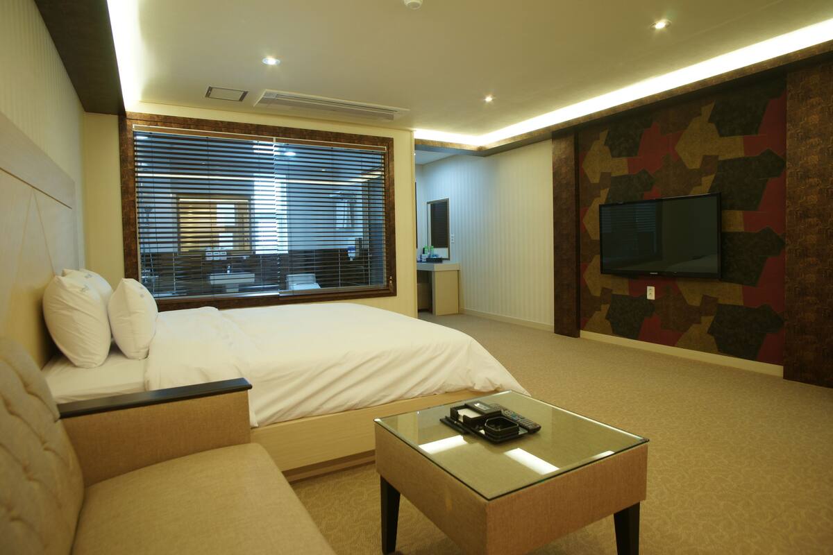 A hotel room with a bed, a glass coffee table, a TV mounted on the wall, and a window with blinds.