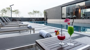 A rooftop pool area with lounge chairs, a table with drinks, and a modern building in the background. - Geno Hotel (Shah Alam)