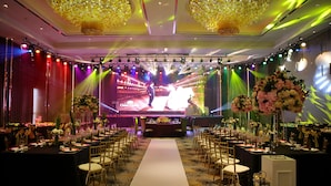 A banquet hall with a long white aisle, tables set for dining, and a stage with a large screen and lighting. - Geno Hotel (Shah Alam)