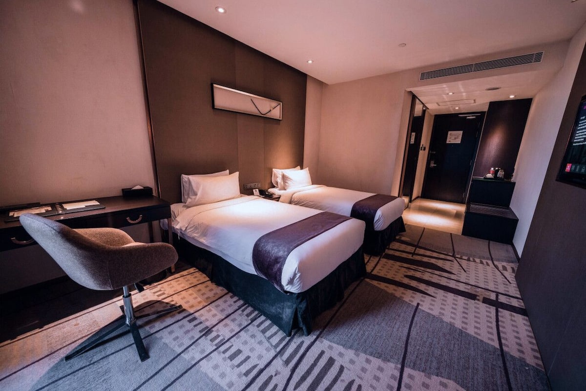 Hotellihuone, jossa on kaksi sänkyä, työpöytä, tuoli ja suuri peili.