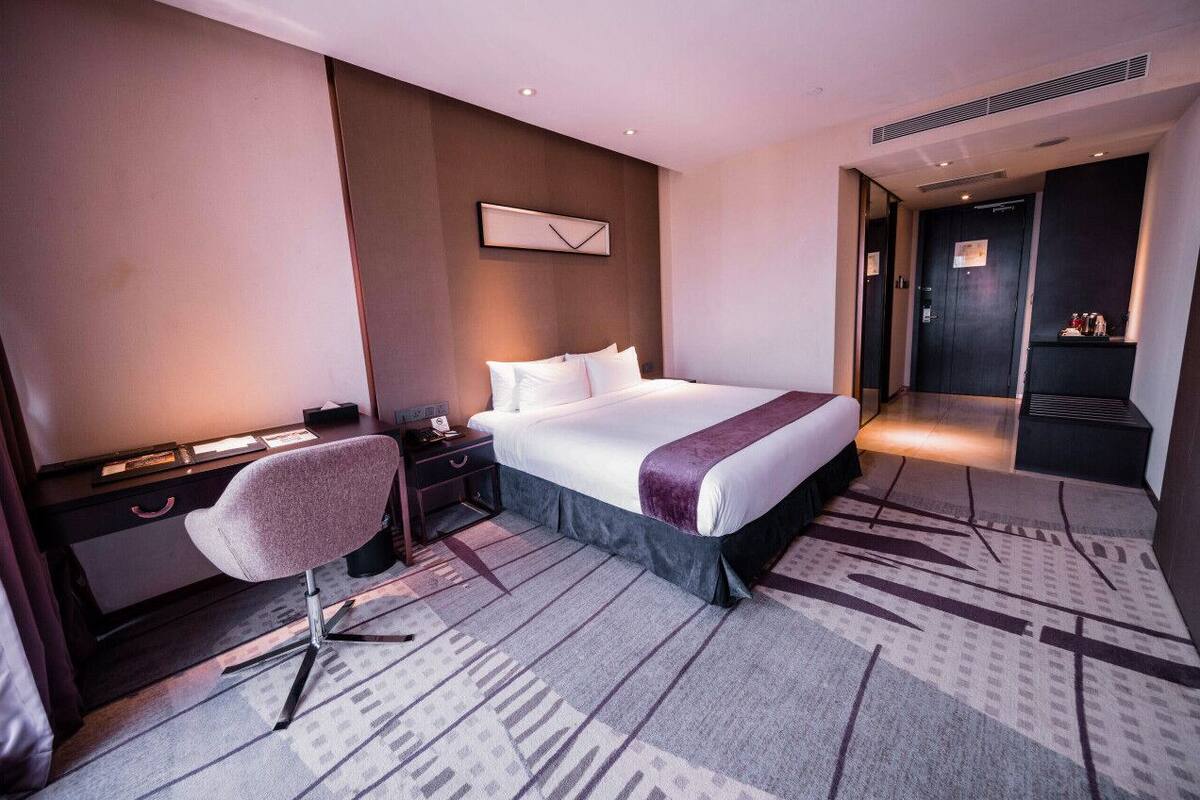 Moderni hotellihuone, jossa on suuri sänky, työpöytä tuolilla, yöpöytä ja seinälle kiinnitetty kello.