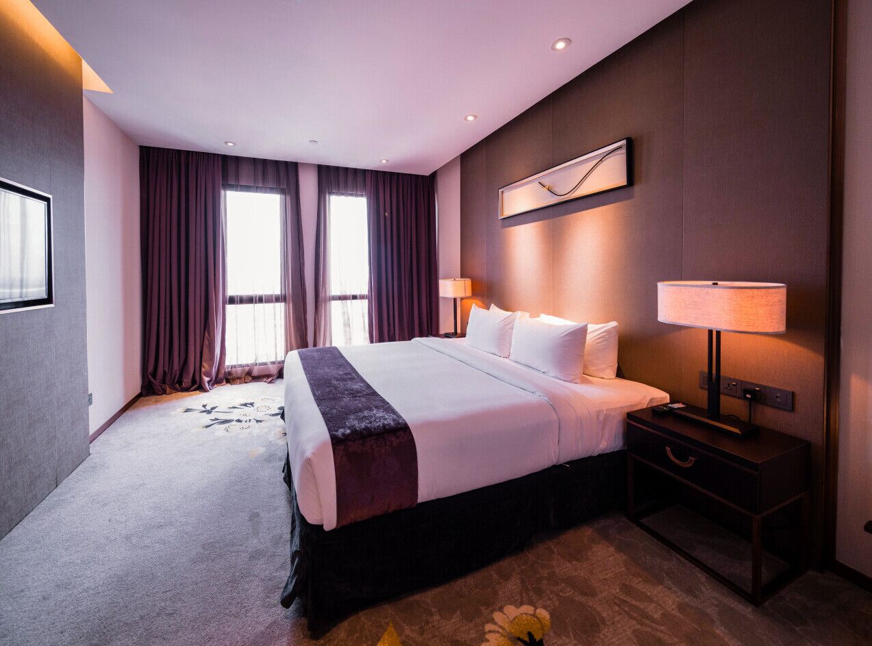 A hotel room with a large bed, a nightstand with a lamp, and a window with curtains.
