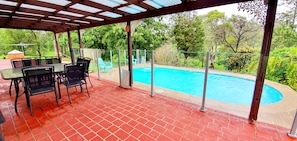 An outdoor patio with a pool, covered seating area, and a view of the surrounding greenery.