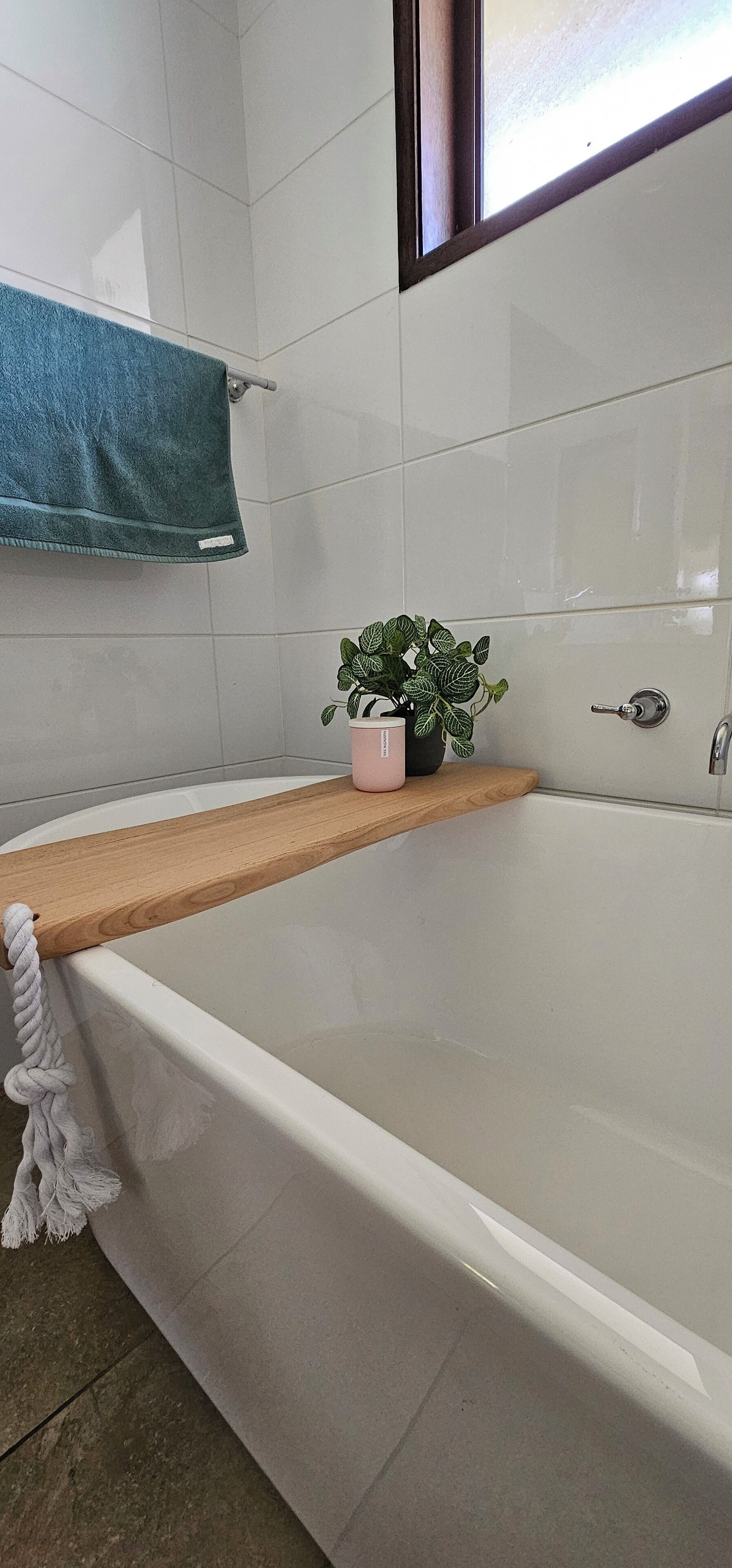 Une salle de bain avec une baignoire blanche, une étagère en bois sur laquelle est posée une plante en pot, et une serviette bleue suspendue à un porte-serviettes.