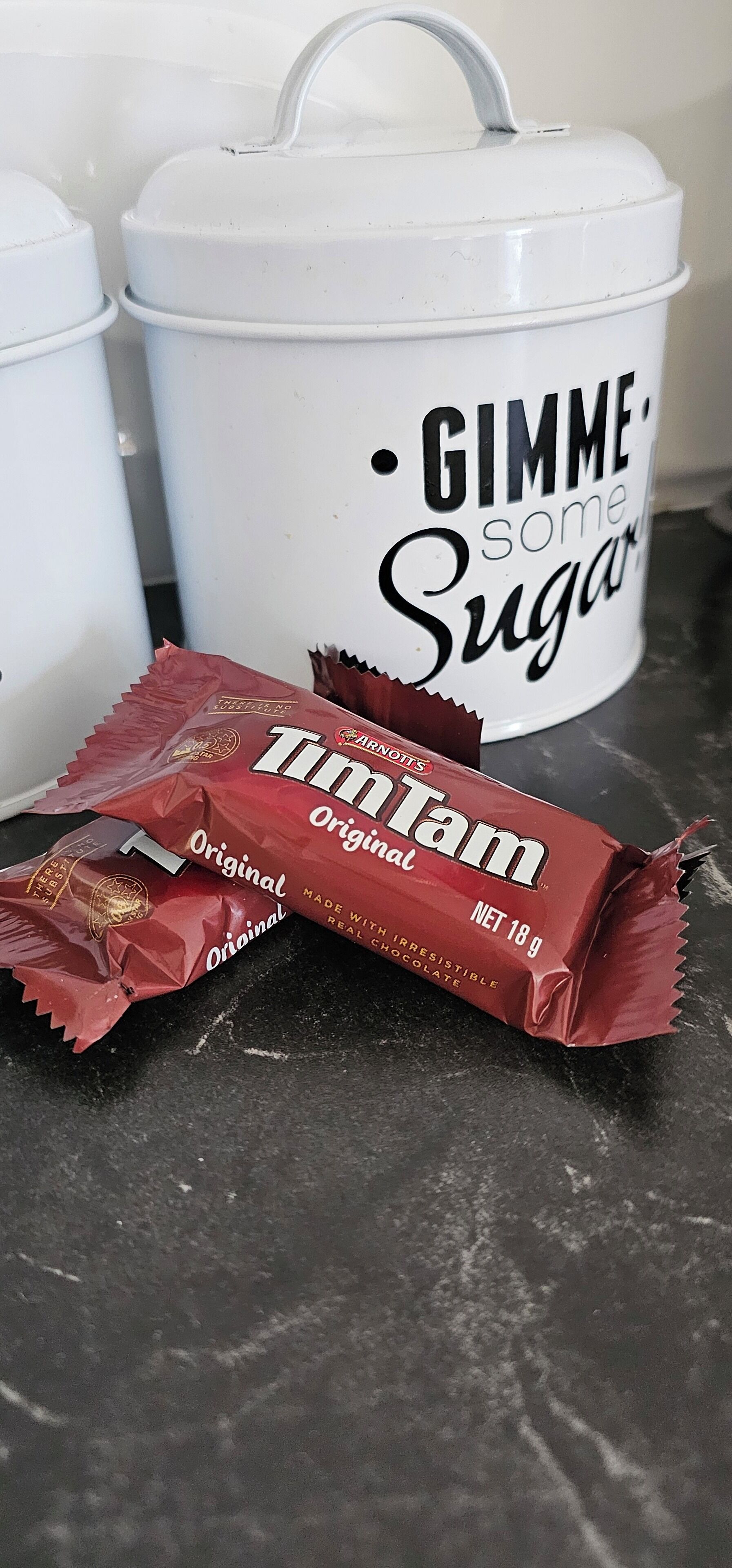 A white container labeled "GIMME some Sugar" and a red Original Tam candy wrapper on a dark surface.