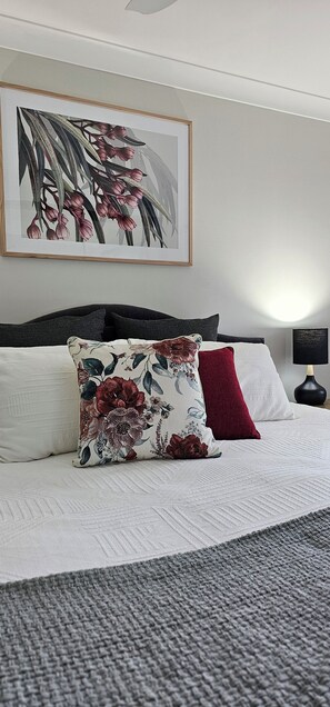 A neatly made bed with a white and gray quilt, a floral pillow, and a red pillow. A framed floral artwork hangs above the bed. - Serendipity On Barrington (Barrington)