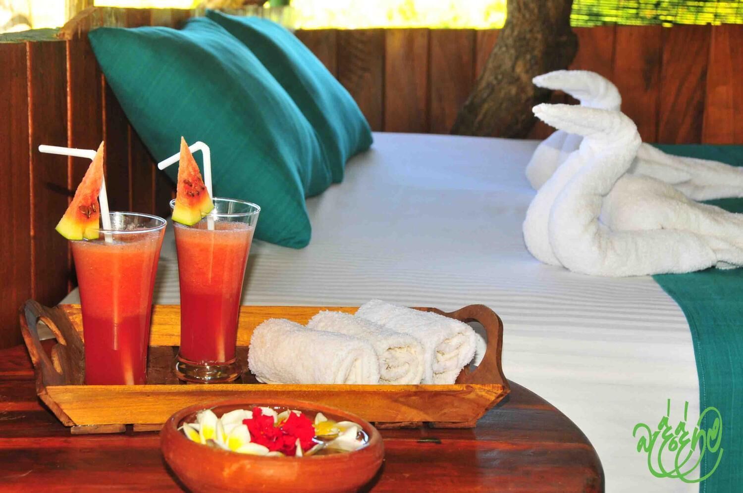 A bed with a green pillow, a tray with two drinks, a bowl of fruit, and a towel.