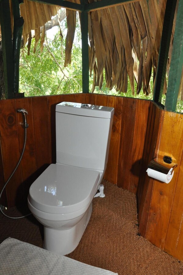 A wooden bathroom with a white toilet, a shower, and a view of greenery outside.