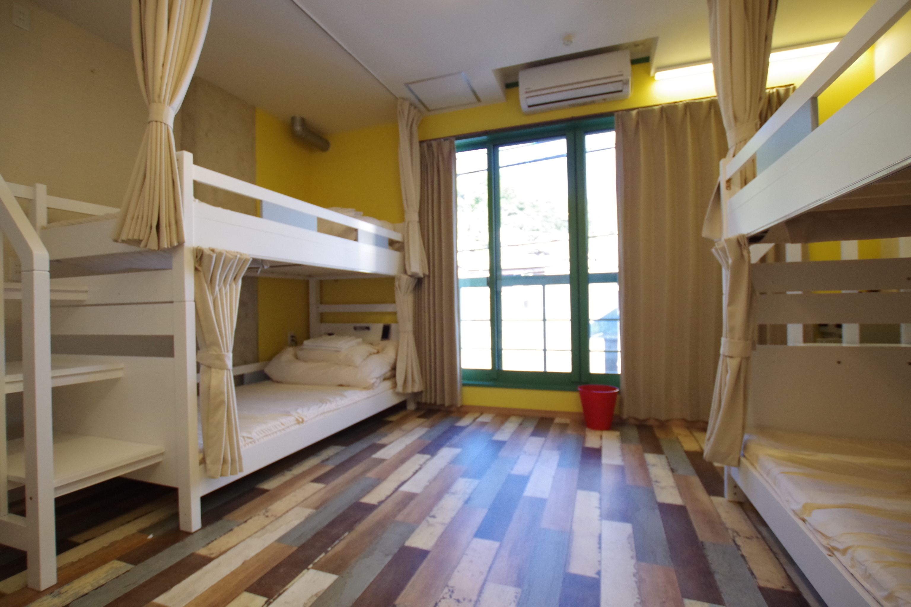 A room with bunk beds, a window with curtains, a red bucket, and an air conditioning unit.