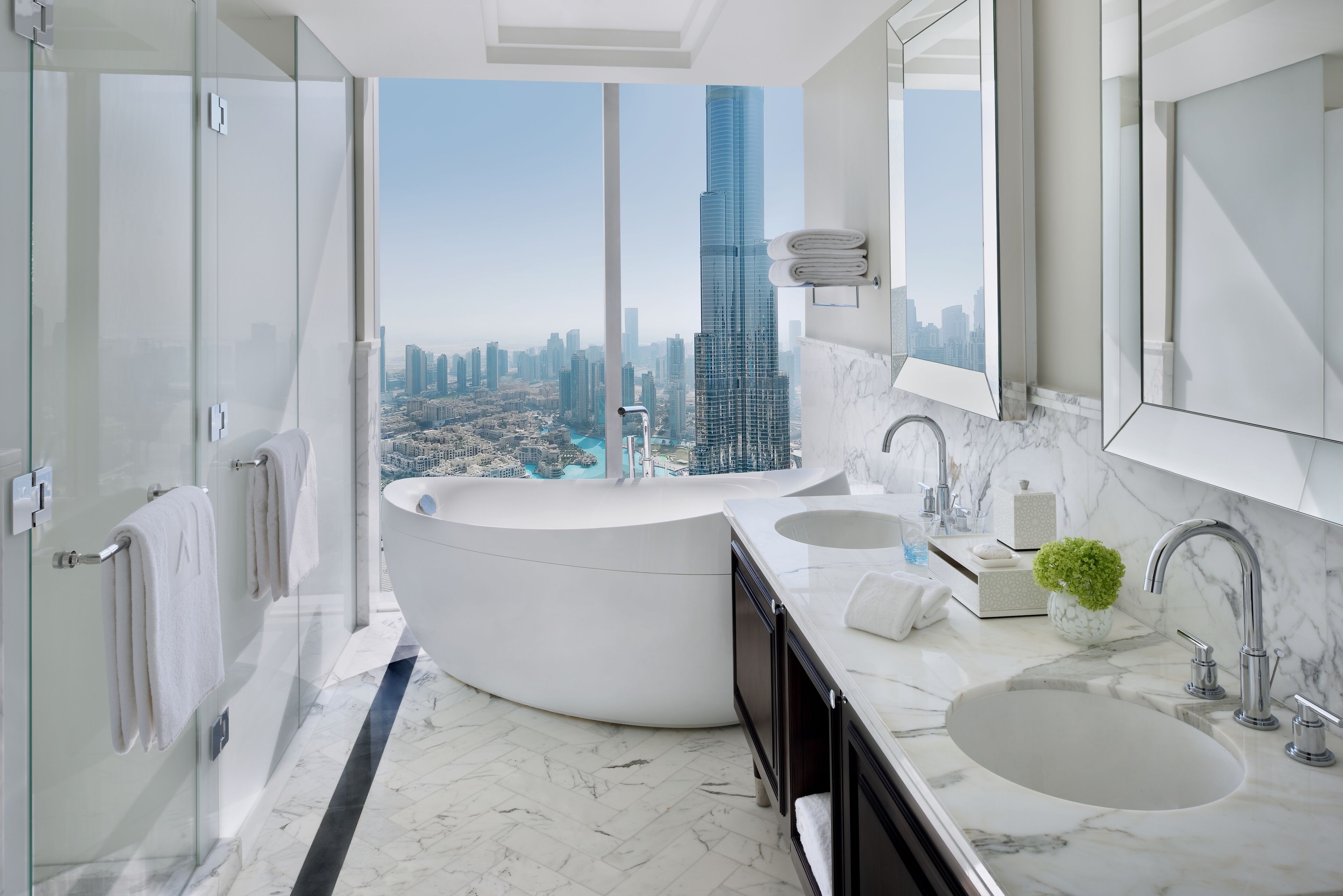 A modern bathroom with a large bathtub, two sinks, and a large mirror. It features a city view with a tall skyscraper.