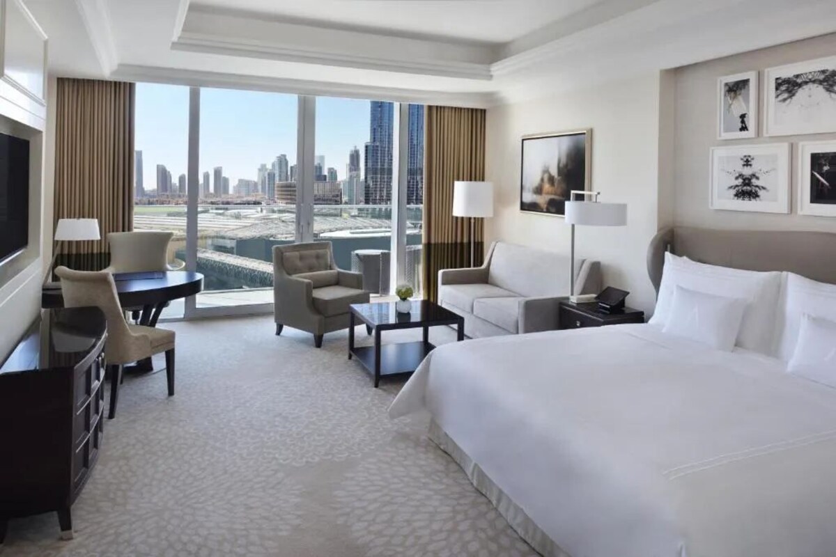 Habitación de hotel con una cama grande, un escritorio, una silla, un sofá y una televisión. Amplias ventanas ofrecen vistas a la ciudad.