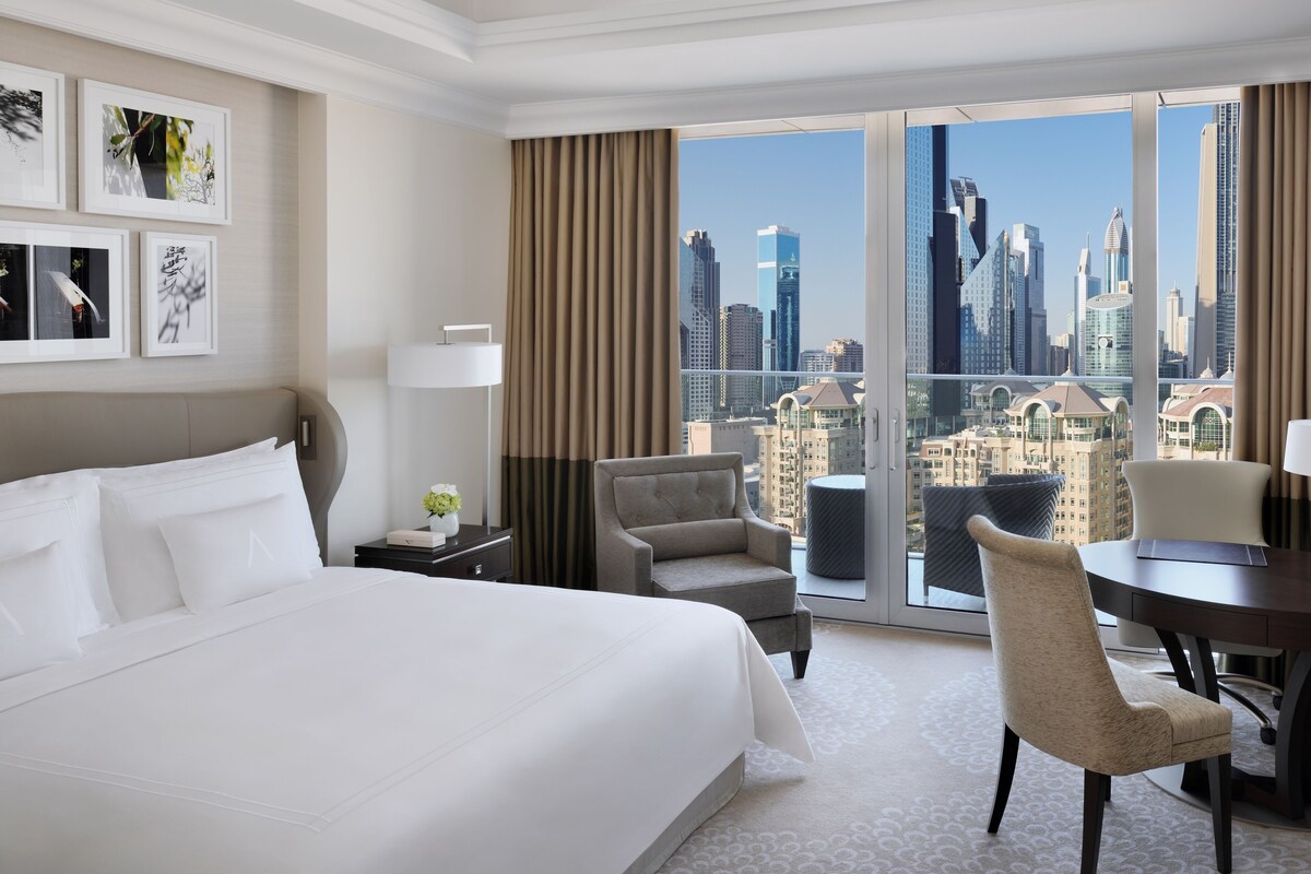 Habitación de hotel con una cama grande, un escritorio con silla, un sofá y vistas a la silueta urbana.