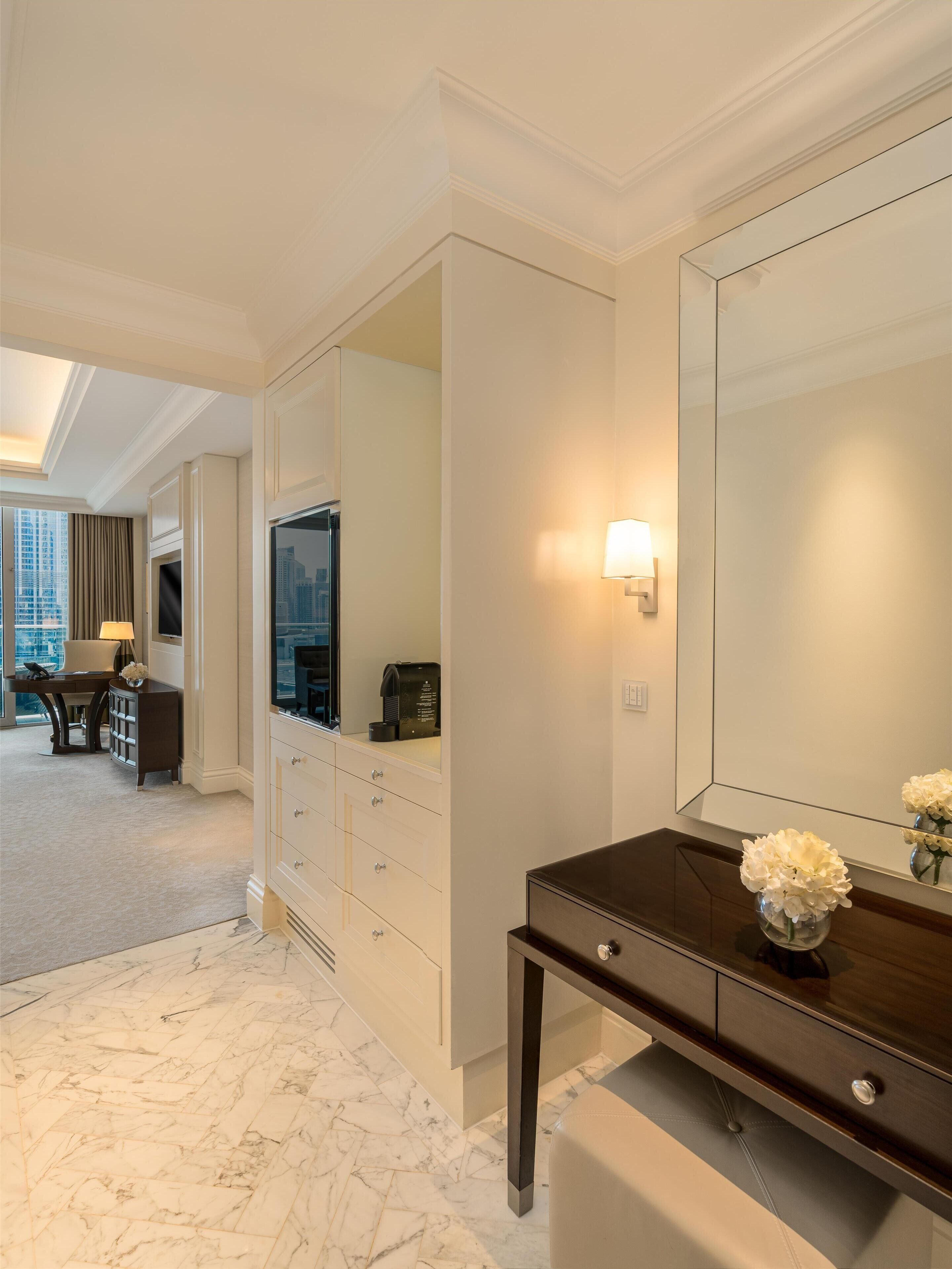 A hotel room with a large mirror, a dark wooden console table with a vase of flowers, a flat-screen TV, and a coffee machine.