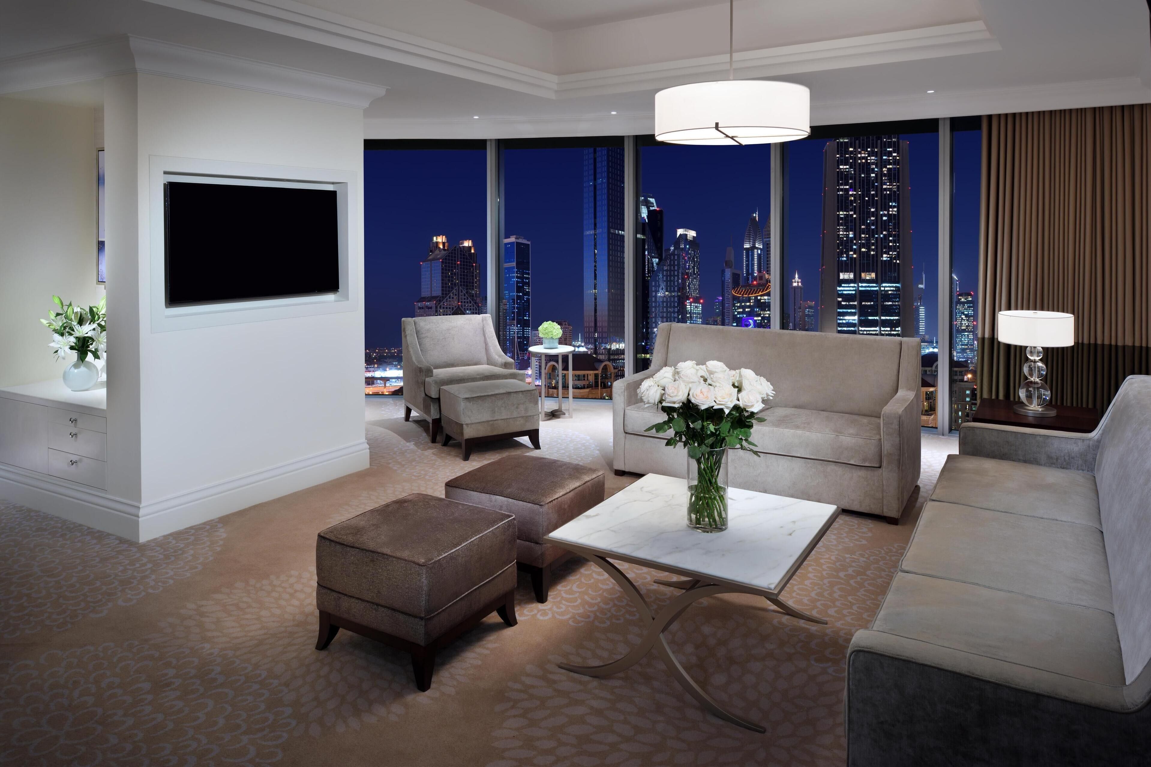 Een moderne hotelkamer met een groot raam en uitzicht op de stadshemel, een flatscreen televisie aan de muur, een bank, twee fauteuils, een bijzettafeltje en een vaas met bloemen.