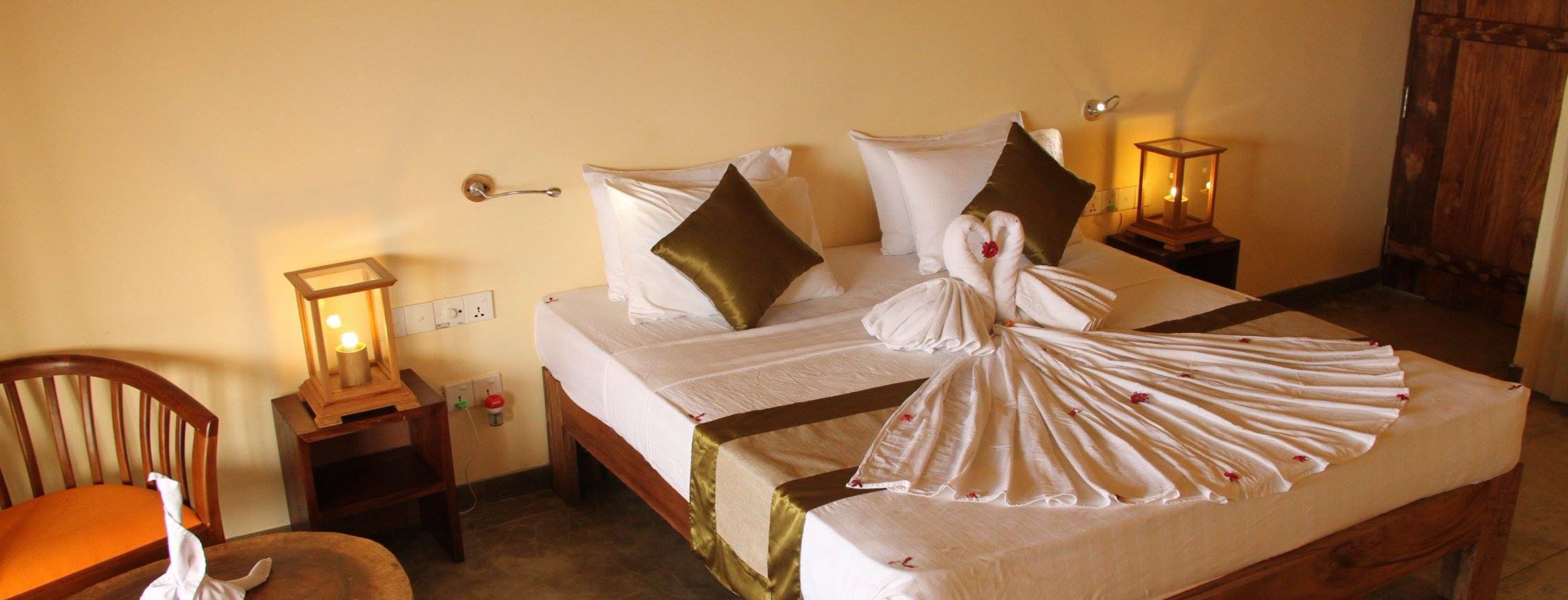 Een netjes opgemaakt bed met witte lakens, een bruine satijnen bedspreider en een handdoek gevouwen in de vorm van een hart. Er staan twee bedlampjes, een houten nachtkastje en een stoel met kussen.