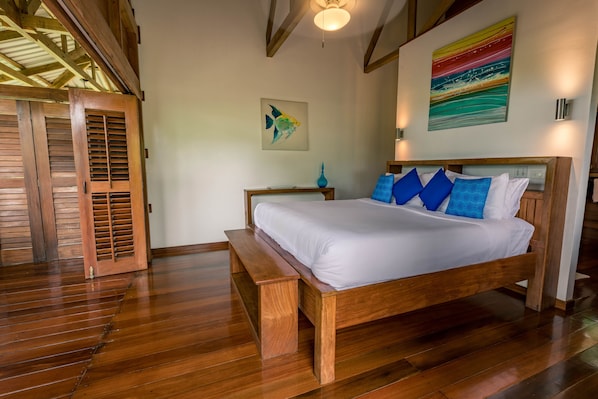 A wooden bed with white bedding, a bench, and a small table with a blue vase. There are two paintings on the walls and wooden doors with louvered windows.