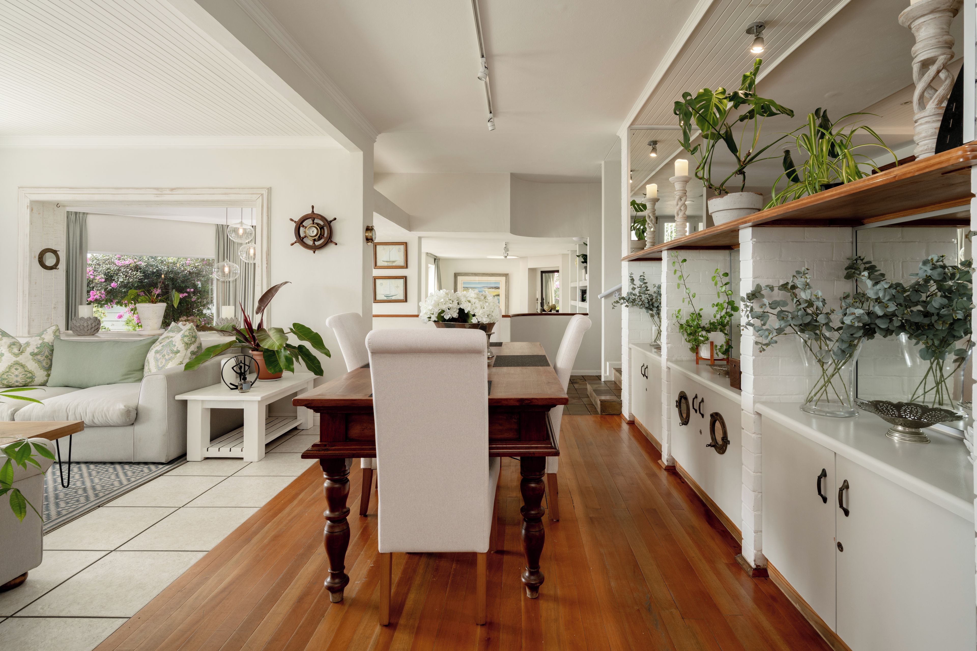 A spacious living room with a wooden dining table, white sofas, and a variety of plants.