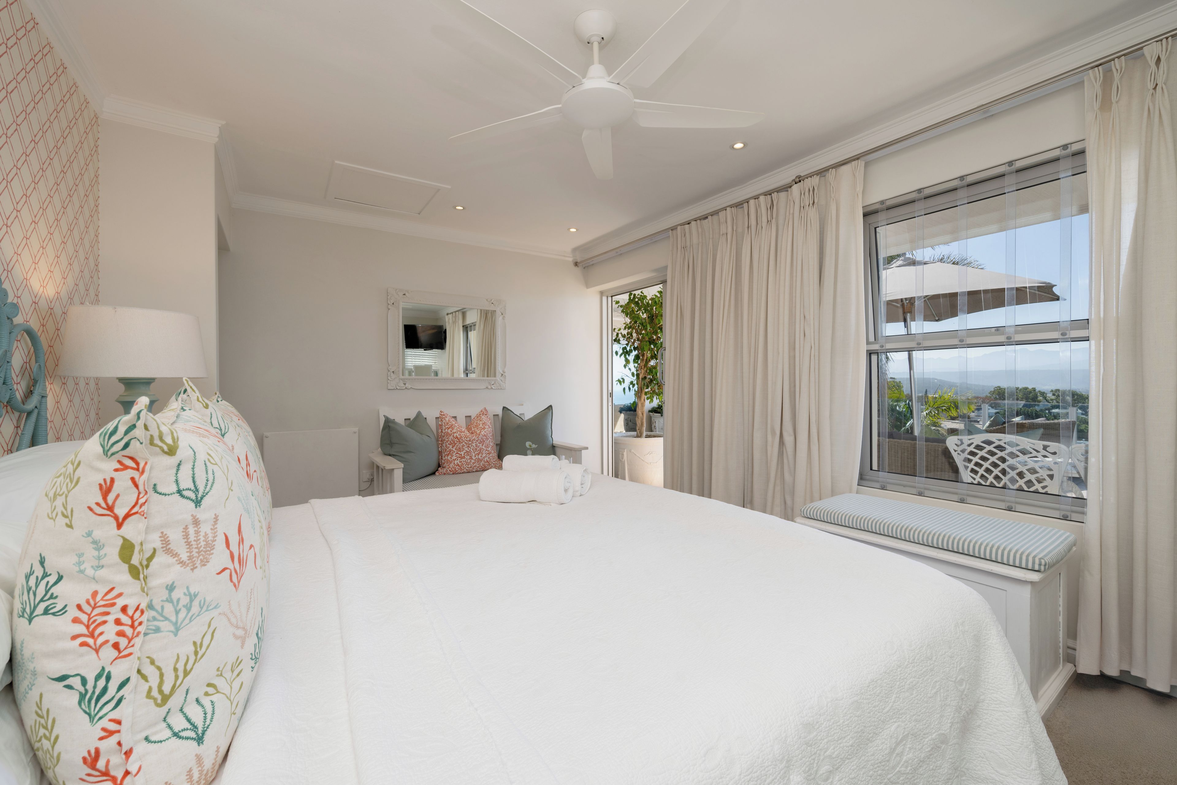 Une chambre d’hôtel avec un grand lit, un ventilateur de plafond, une fenêtre avec des rideaux, une vue sur l’extérieur et une petite table avec une lampe.