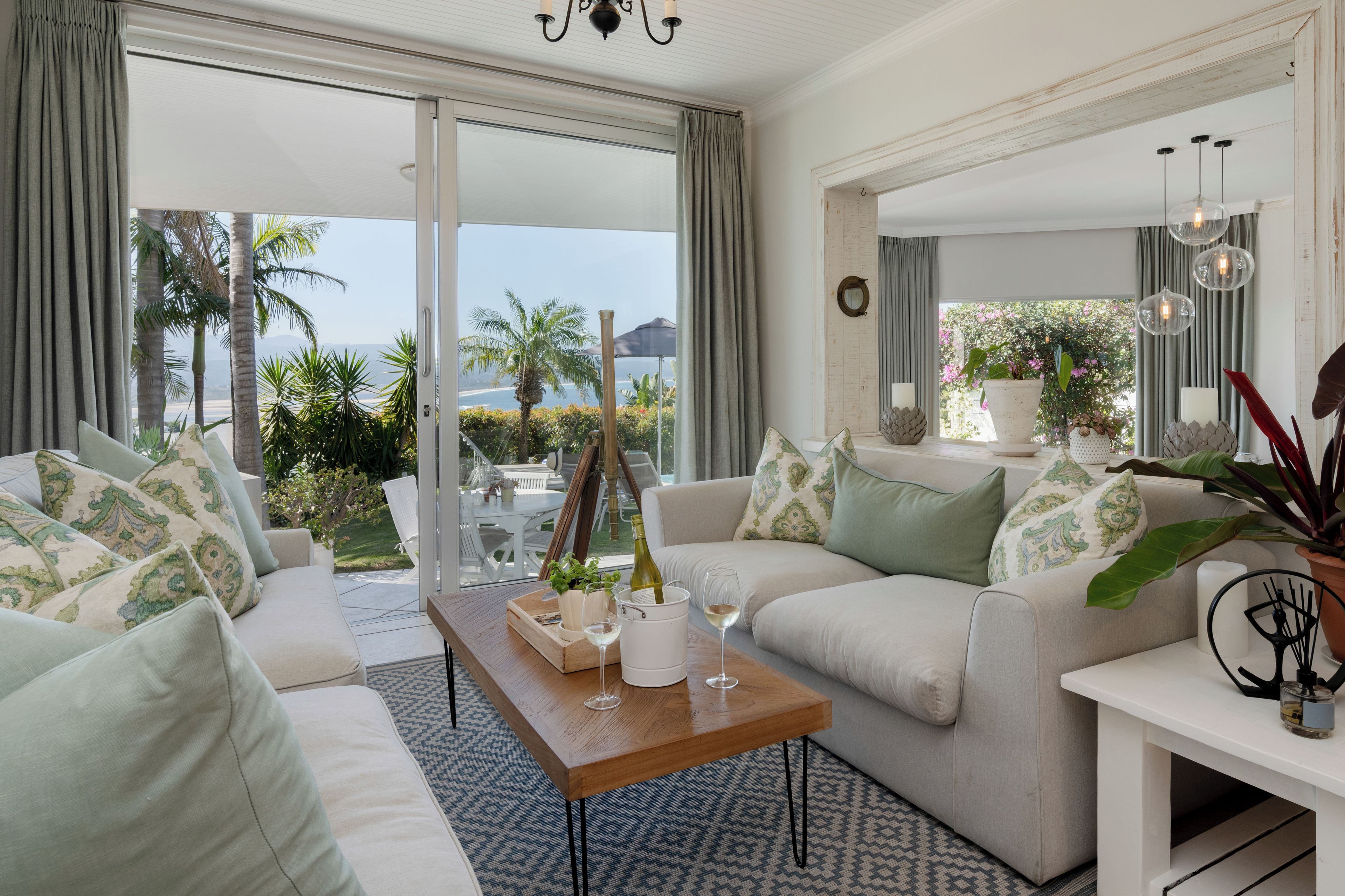 A modern living room with a view of the ocean, featuring two sofas, a coffee table with a bottle and glasses, and a potted plant.