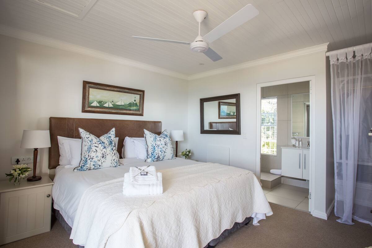 A neatly arranged bedroom with a large bed, a ceiling fan, a bedside table with a lamp, and a framed picture on the wall.