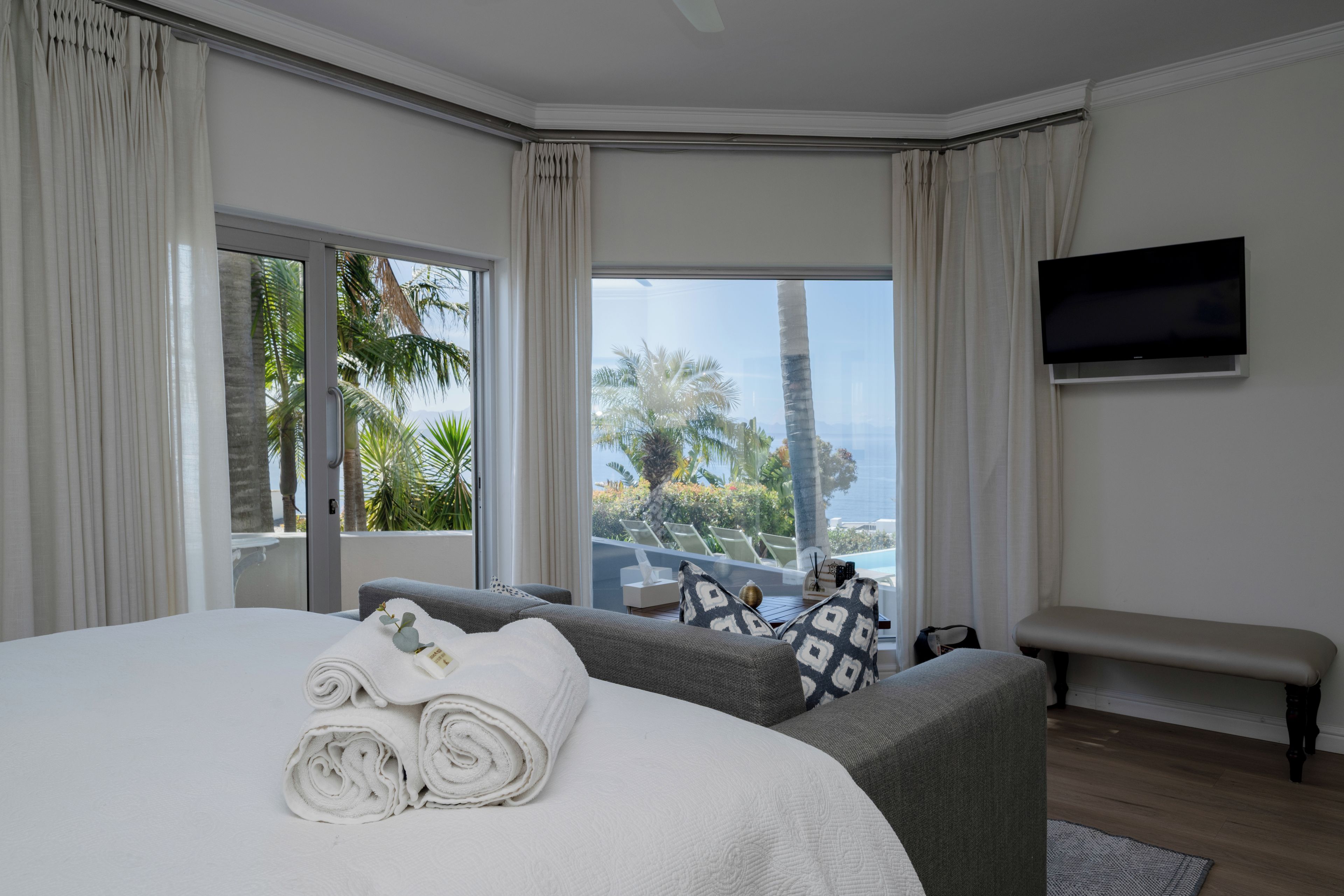 Une chambre dotée d’un grand lit, d’un banc, d’un téléviseur à écran plat et offrant une vue sur des palmiers et une piscine.