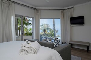 Un dormitorio con una cama grande, una banca, un televisor de pantalla plana y vistas a palmeras y una piscina.