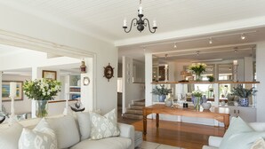 Amplia sala de estar con un sofá grande, una mesa de centro de madera y una araña. Se ve una escalera que lleva a un nivel superior, y una zona de cocina con un espejo grande y elementos decorativos.